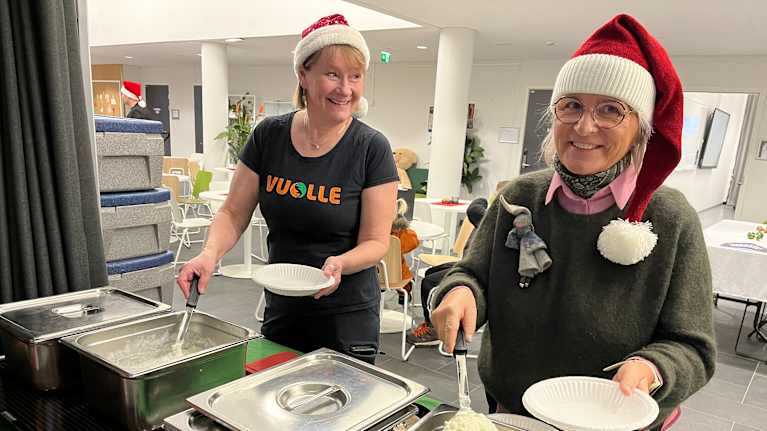 Kaksi tonttulakkipäistä ihmistä nostelee puuroa lautaselle.