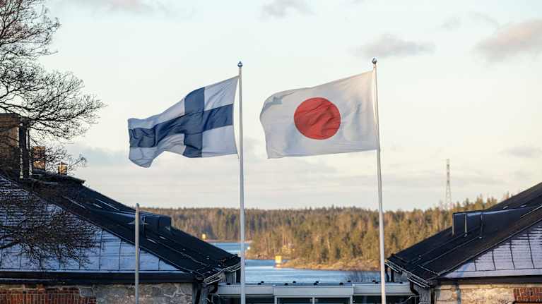 Kuvassa Suomen ja Japanin liput liehuvat vierekkäin ulkona. Taustalla näkyy metsäinen maisema.
