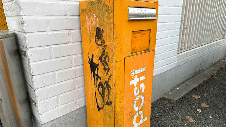 Postin keltainen, töhritty kirjelaatikko valkotiilisen rakennuksen seinustalla.