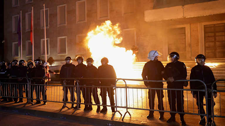 Poliisit ovat asettuneet riviin rakennuksen edustalle mellakka-aidan taakse. Heidän takanaan räjähtää.