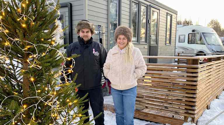 Mies ja nainen seisovat talvivaatteissa kameralle hymyillen. Etualalla joulukuusi, taustalla mökkivaunu ja asuntoauto.