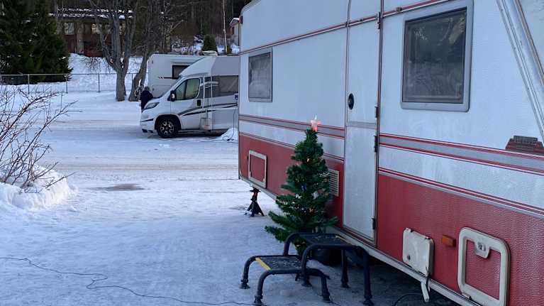 Talvinen leirintäalue, jossa kaksi matkailuautoa. Etualalla punavalkoisen matkailuauton vieressä on pieni joulukuusi. Luminen maa ja taustalla puita.