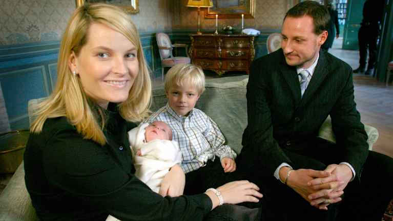 Mustiin pukeutuneet nainen ja mies. Naisen sylissä vauva ja pariskunnan välissä sohvalla vähän isompi poika.