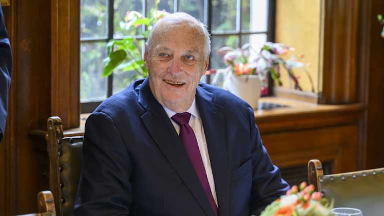 Kuningas Harald istuu pöydän ääressä. Taustalla ikkuna.