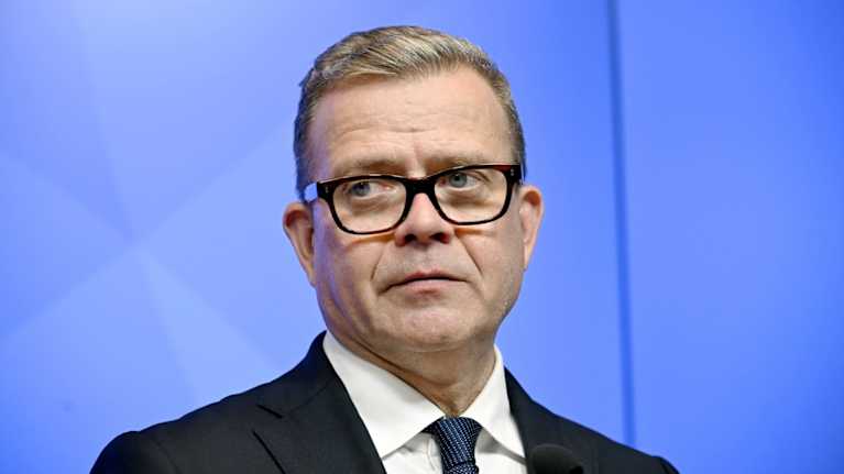 Pääministeri Petteri Orpo katsoo kohti median edustajia. Taustalla sininen seinämä.