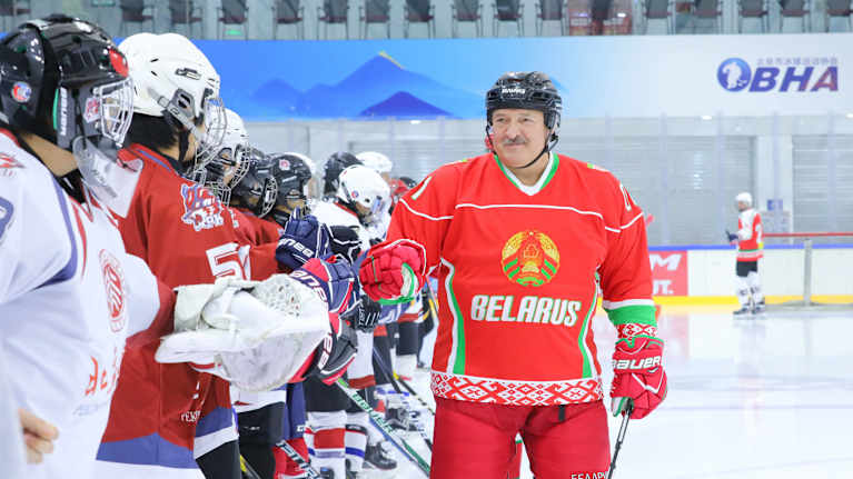 Viiksekäs mies Valko-Venäjän pelipaidassa tekee nyrkkitervehdyksiä rivissä seisovien jääkiekon pelaajien kanssa.