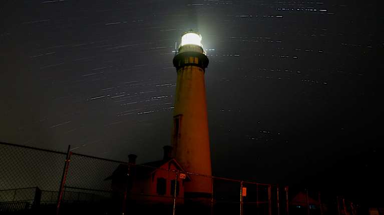 Tähdenlennot muodostavat valojuovia majakan takana tummalla yötaivaalla.