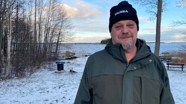 Mies seisoo lumisella kentällä meren rannalla ja katsoo kameraan.