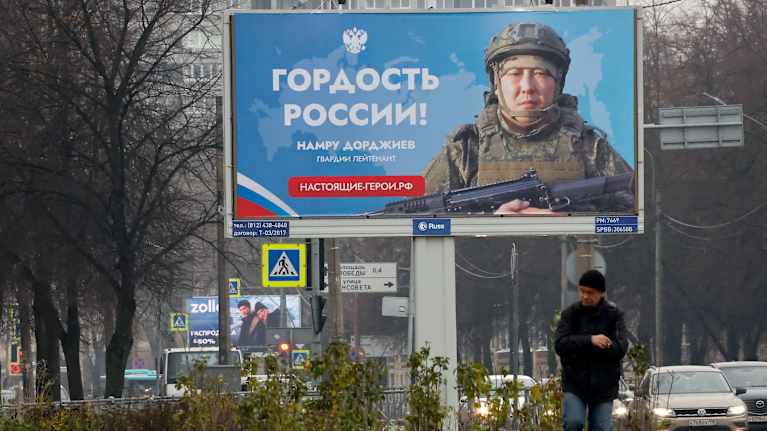 Mies ulkoiluttaa kissaa kadun viereisessä puistossa. Taustalla on suurikokoinen juliste, jossa on kuva sotilaasta sekä teksti: "Venäjän ylpeys!".