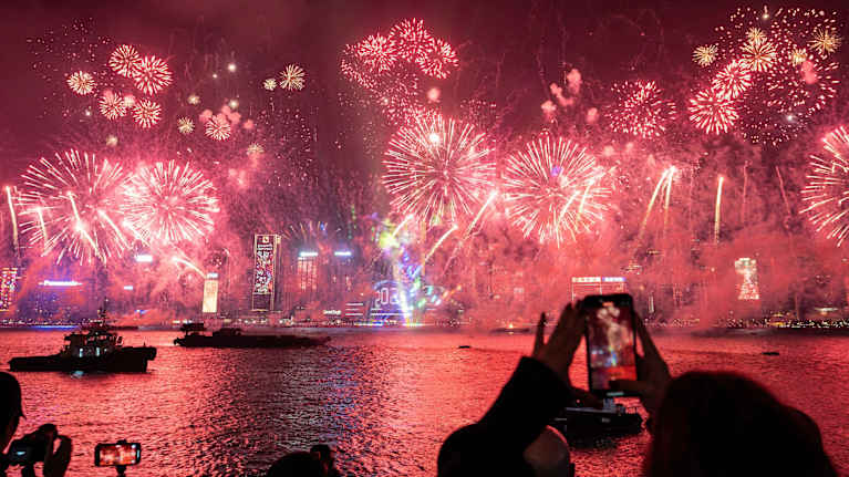 Ilotulitteet räiskyvät taivaalla Hongkongin satamassa. Etualalla joku kuvaa tulitusta kännykällään.