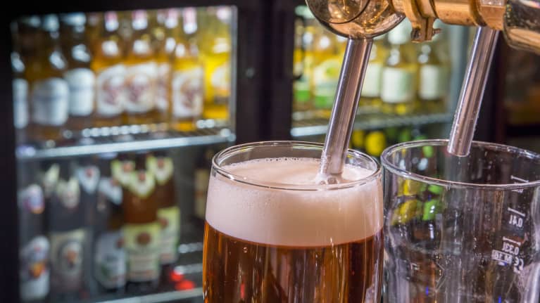 Kaksi lasia, joissa toisessa on olutta. Taustalla näkyy olutpulloja kaapissa.