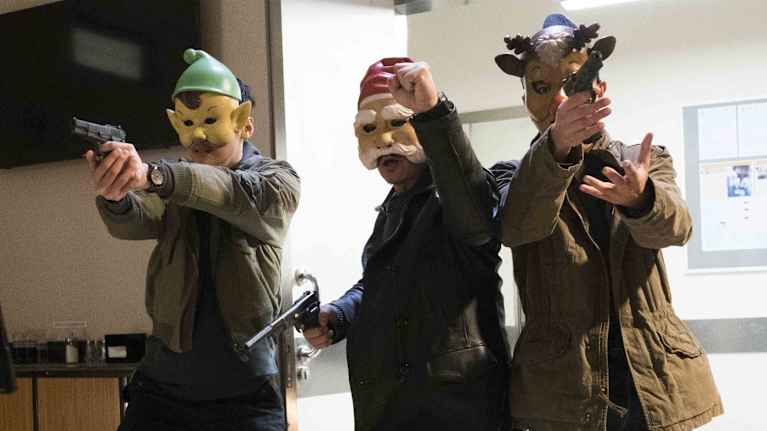 Tre män med masker som föreställer en julgubbe, tomte och en ren rusar in genom en dörr med dragna vapen.