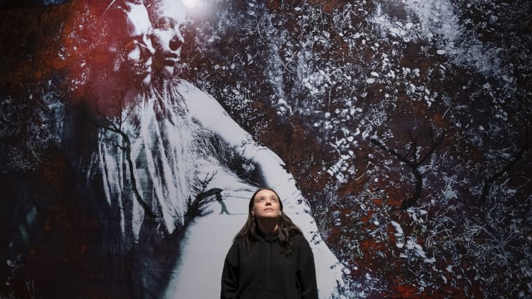 Lada Suomenrinne seisoo taideteoksensa edessä ja katsoo yläviistoon. 