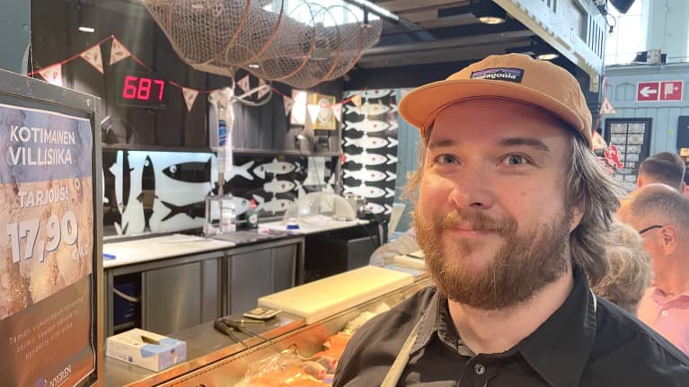 Parrakas ja aavistuksen pitkähiuksinen mies oranssi lippis päässä katsoo kameraan hymyillen. Taustalla näkyy Tamperen kauppahallin asiakkaita ja kalatiski.