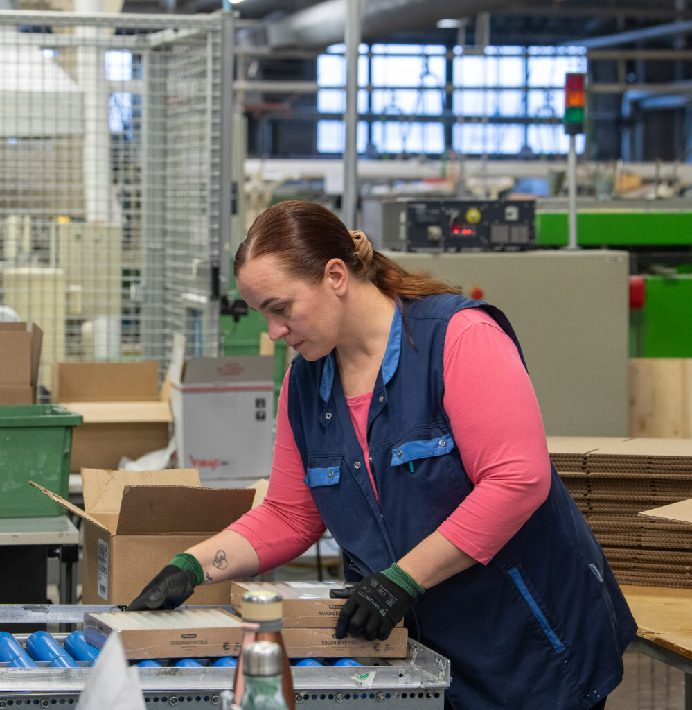 Henkilö pitelee ja katsoo pahvisia pakkauksia, joissa on kynttilöitä. Pakkaukset ovat rullien päällä. Takana näkyy tehdassalin putkia, koneita ja laitteita.