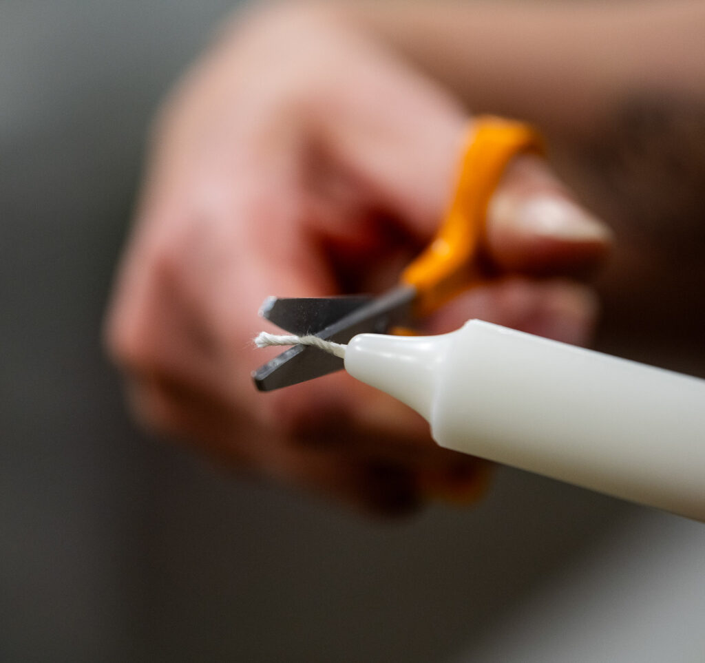 Kruunukynttilän sydäntä leikataan lyhemmäksi saksilla. Leikkaajan käsi näkyy lähikuvassa.