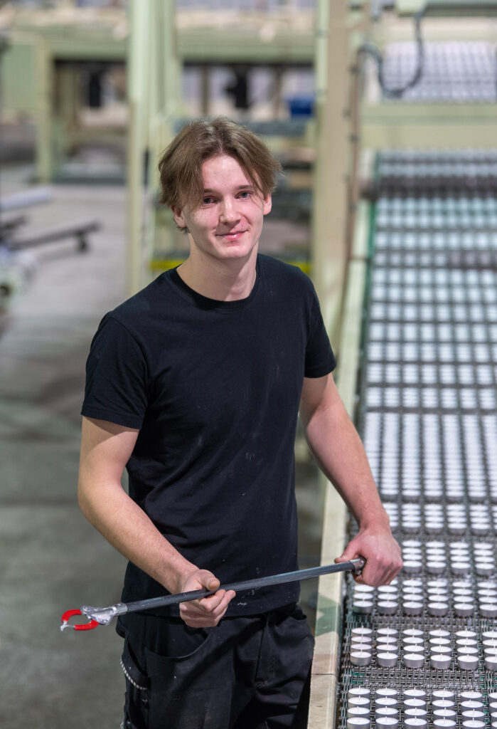 Henkilö seisoo tehtaan kynttilälinjaston vieressä. Hän katsoo kameraan ja käsissään hänellä on pitkulainen työkalu.