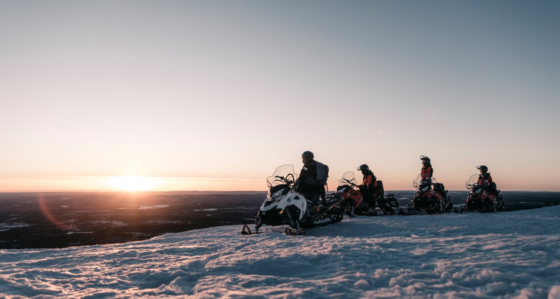 Neljä moottorikelkkailijaa ovat pysähtyneenä tunturin huipulla. Auringon lasku näkyy taustalla.