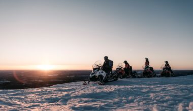 Neljä moottorikelkkailijaa ovat pysähtyneenä tunturin huipulla. Auringon lasku näkyy taustalla.
