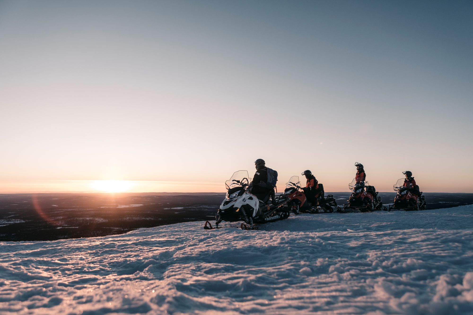 Neljä moottorikelkkailijaa ovat pysähtyneenä tunturin huipulla. Auringon lasku näkyy taustalla.