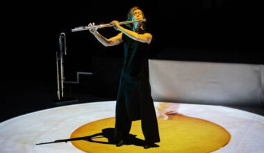 Kuvassa on Claire Chaise soittamassa huilua seisten ja hänellä on musta hihaton musta mekko sekä pitkät housut. Hän seisoo keltaisen, pyöreän maton päällä ja sen ympärillä on valkoinen ympärillä. Taustalla on mustaa.