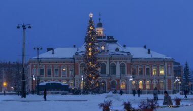 Suuri joulukuusi koristeltuna valoilla ja tähdellä latvassa seisoo lumisella torilla iltahämärässä. Taustalla näkyy näyttävä, historiallinen rakennus, jonka julkisivu on valaistu."
