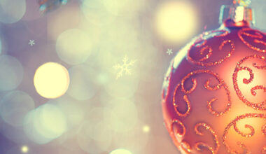 Punasävyinen kimalteleva joulupallo ja taustalla valojen kimallusta ja lumihiutaleita.