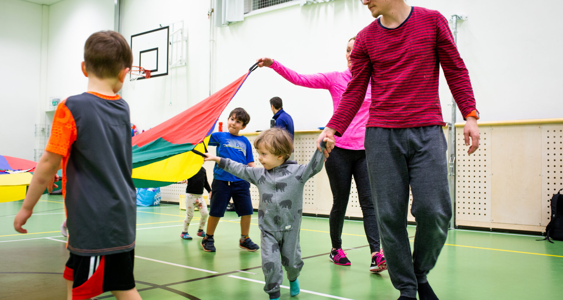 Lapsia ja vanhempia liikkumassa leikkivarjon kanssa liikuntasalissa.