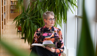 Henkilö istuu kirjastossa kirjahyllyjen ja viherkasvien ympäröimänä, katsoo ulos ikkunasta kirja sylissään.