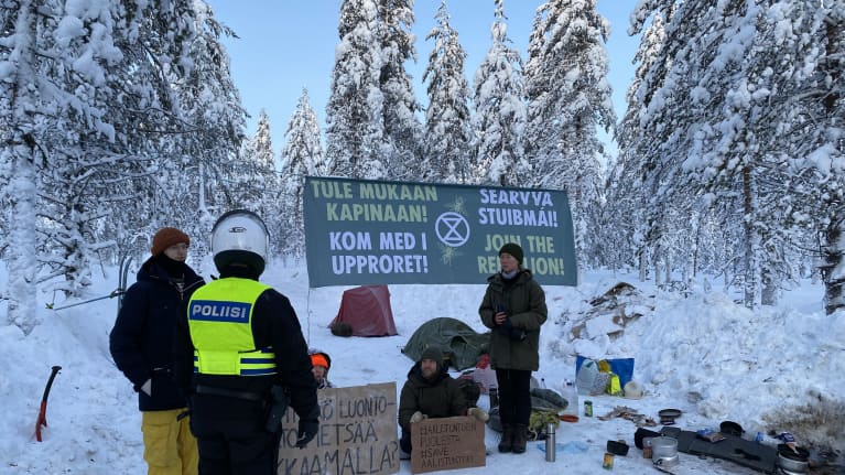 Neljä mielenosoittajaa seisoo kylttiensä edessä lumisessa metsämaisemassa, edessä selin poliisi.