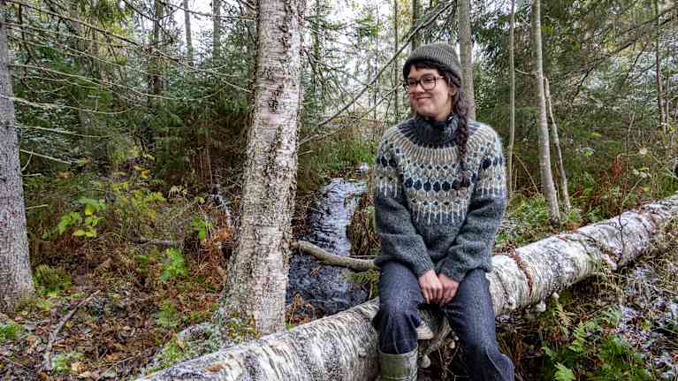 Väitöskirjatutkija Veera Saari istuu suojellun puron varrella kaatuneen puunrungon päällä.