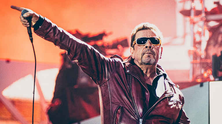 Pate Mustajärvi puolilähikuvassa punaisessa nahkatakissa ja aurinkolaseissa. Hänen oikea kätensä on kohotettuna sivulla etusormi pystyssä, ja hän pitelee siinä mikrofonia.