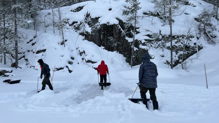 Kolme ihmistä kolaa lunta järveltä kallioiselle rannalle isoon kinokseen.