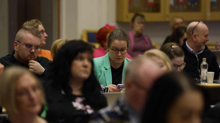 Vihreätakkinen nainen selaa puhelinta aluevaltuuston kokouksessa muiden kokoustajien keskellä