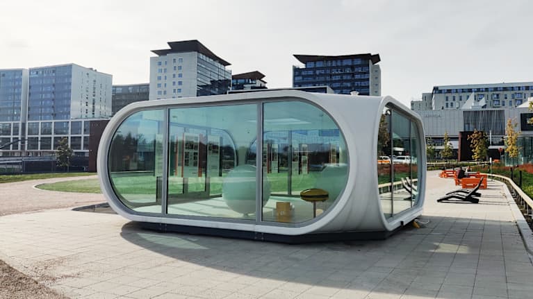 Utopistinen, pehmeäkulmainen ja matala lujitemuovista valmistettu Venturo-talo hiekkakentällä kauppakeskus Ainoan edustalla.