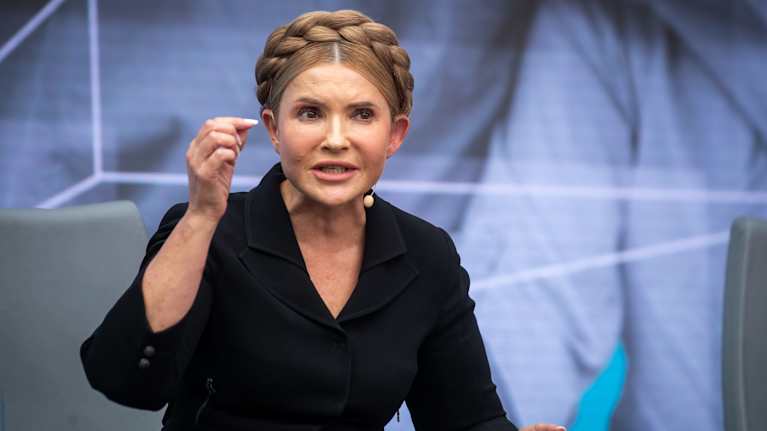 Kuvassa on Ukrainan entinen pääministeri Tymoshenko puhumassa mahdollisesti tilaisuudessa tai haastattelussa. Hänellä on yllään musta asu ja hiukset on laitettu kauniille letille pään ympärille. Taustalla näkyy suurikokoinen kuva tai juliste.