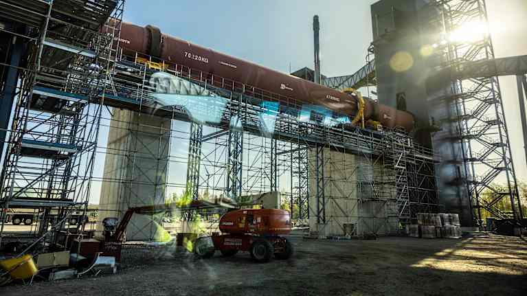Teollisuusrakennus auringonpaisteessa, jossa näkyy suuri sylinterimäinen rakenne ja rakennustelineitä. Etualalla on nosturi ja rakennusmateriaaleja.