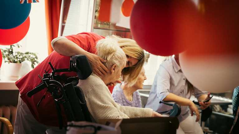 Hoitaja avustaa pyörätuolissa istuvaa naista.