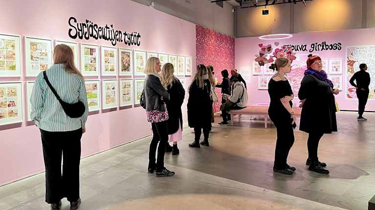 Kuvassa on sarjakuvataidenäyttelyn huone, jossa seinillä näkyy useita taideteoksia. Ihmiset seisovat katsomassa teoksia.