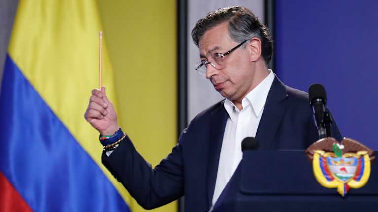 Gustavo Petro pitää oikeassa kädessään lyijykynää opettavaisesti. Hänellä on tumma puku, kauluspaita mutta ei kravattia. Taustalla on Kolumbian lippu.