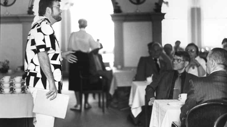 Mustavalkoiseen paitaan pukeutunut mies puhuu pöydässä istuvalle yleisölle.