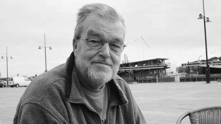 Mies istuu ulkona syyspäivänä, taustalla kaupunkimaisema ja ravintolalaiva.