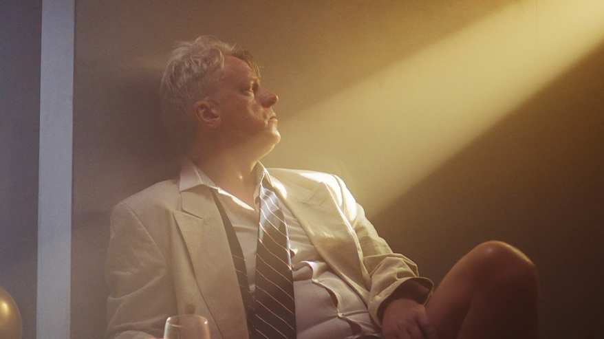 Lavavstettu esityskuva. Mies valkoinen puku päällä, mutta ilman housuja. Katsoo ylös kohti ikkunasta tulevaa valoa. Kädessä lähes tyhjä punaviinilasi. 