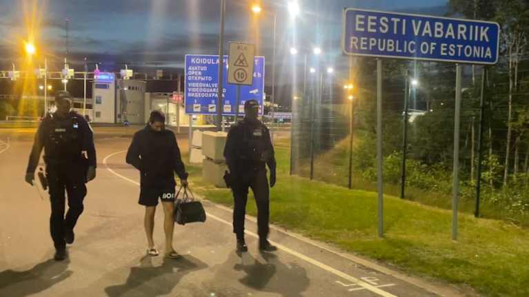 Kaksi miestä Viron poliisin kenttäpuvussa kävelee Viron ja Venäjän välisellä rajanylityspaikalla, välissään kassia kantava mies shortseissa.