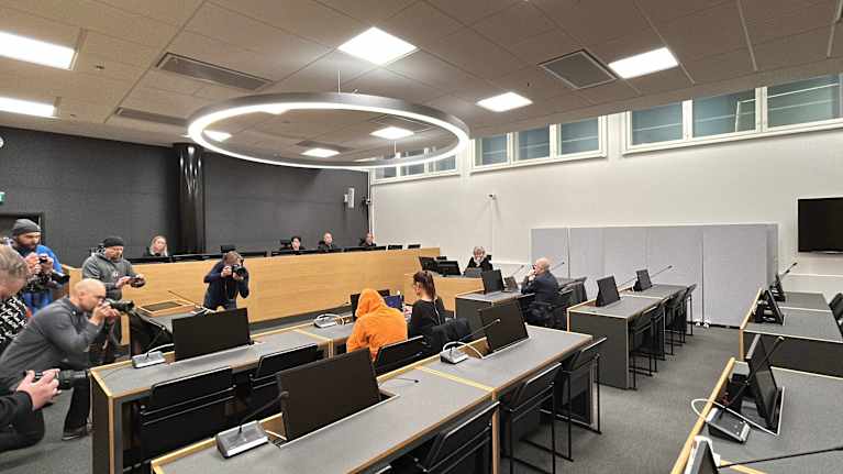 Käräjäoikeuden salissa oranssiin huppariin pukeutunut vastaaja istuu pöydässä asianajajan kanssa. Kuvaajat kuvaavat.