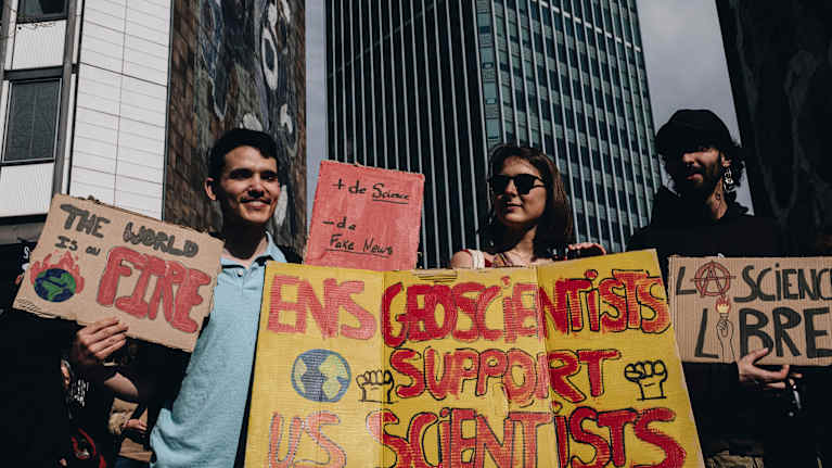 Kolme hymyilevää ihmistä mielenosoituskyltteineen modernien rakennusten edessä auringonpaisteessa.