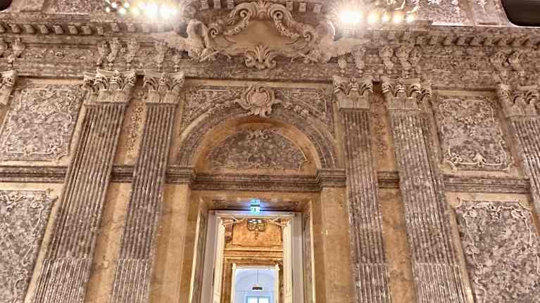 Koristeltua beessinväristä marmoriseinää ja eteenpäin pylväikkökäytävää, jolla näkyy kaksi tummaa hahmoa.