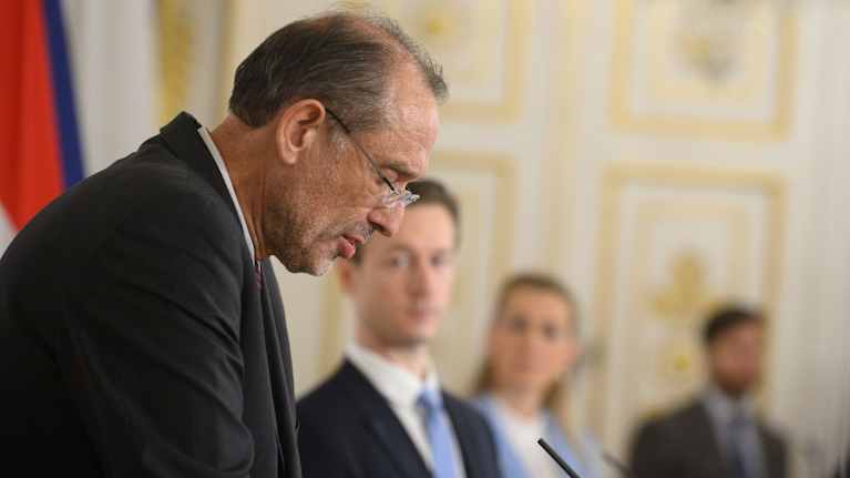 Sivuprofiilissa myöhäiskeski-ikäinen pukumies, taustalla näkyy kolme ihmishahmoa ja kultaista seinää.