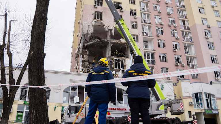 Kaksi henkilöä katsomassa nosturia, joka on asetettu vahingoittuneen rakennuksen eteen. Rakennuksessa on merkittäviä vaurioita useiden kerrosten kohdalla. 