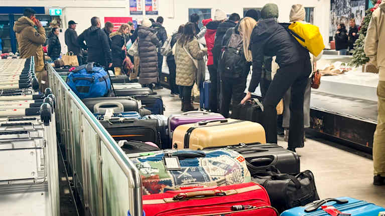 Matkalalukkuja lattialla laukkuhihnan vierellä. Ihmiset odottavat laukkujaan hihnalta.